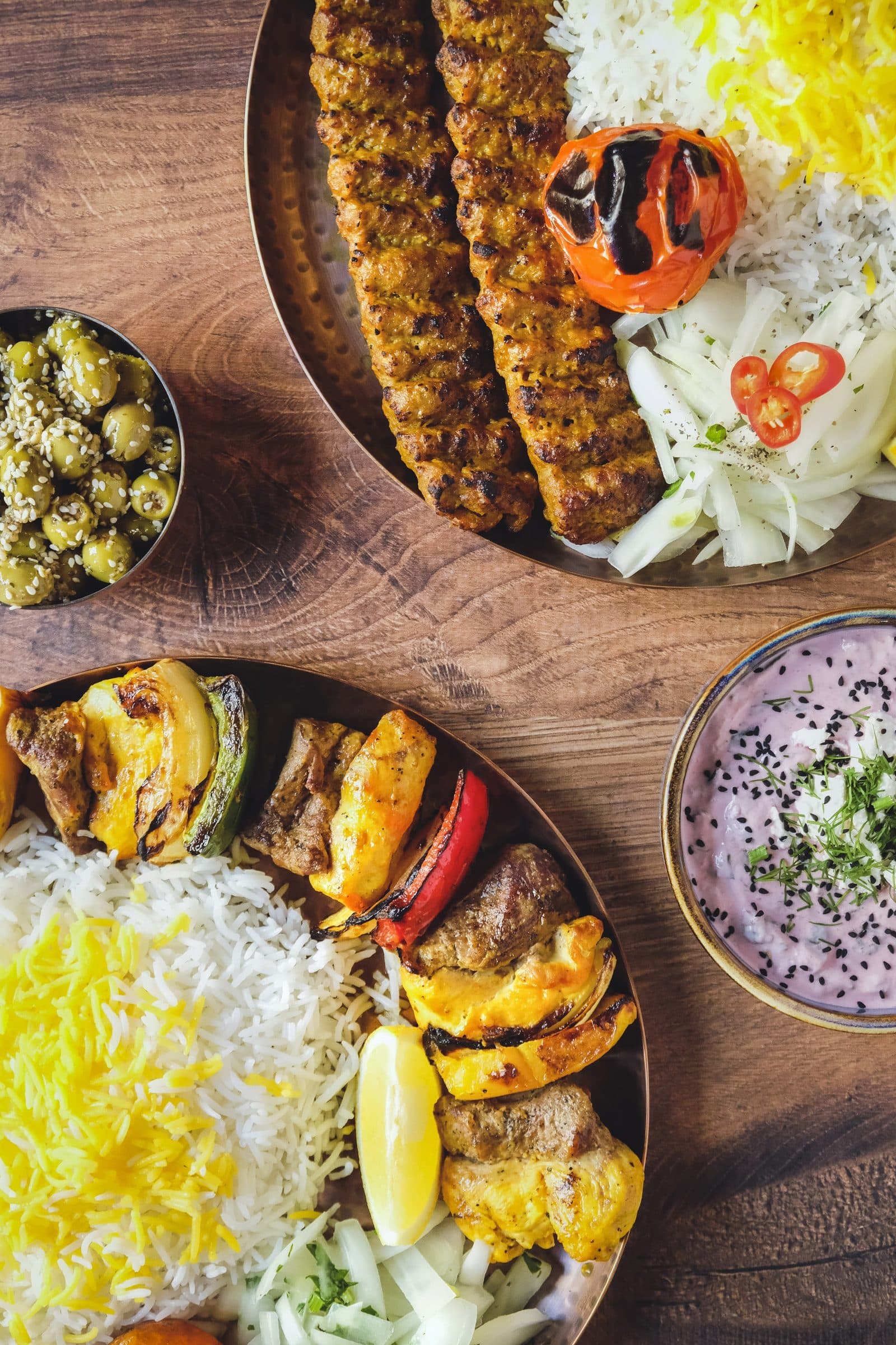 Koubideh kebab on saffron rice with grilled tomato, beside a platter of charcoal-grilled skewers and vegetables